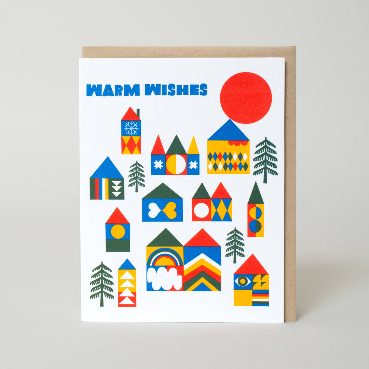 A white greeting card with colorful geometric house illustrations and the words 'WARM WISHES' in blue, shown with a kraft envelope against a light gray background. Warm Wishes Village Letterpress Greeting Card by Lisa Congdon