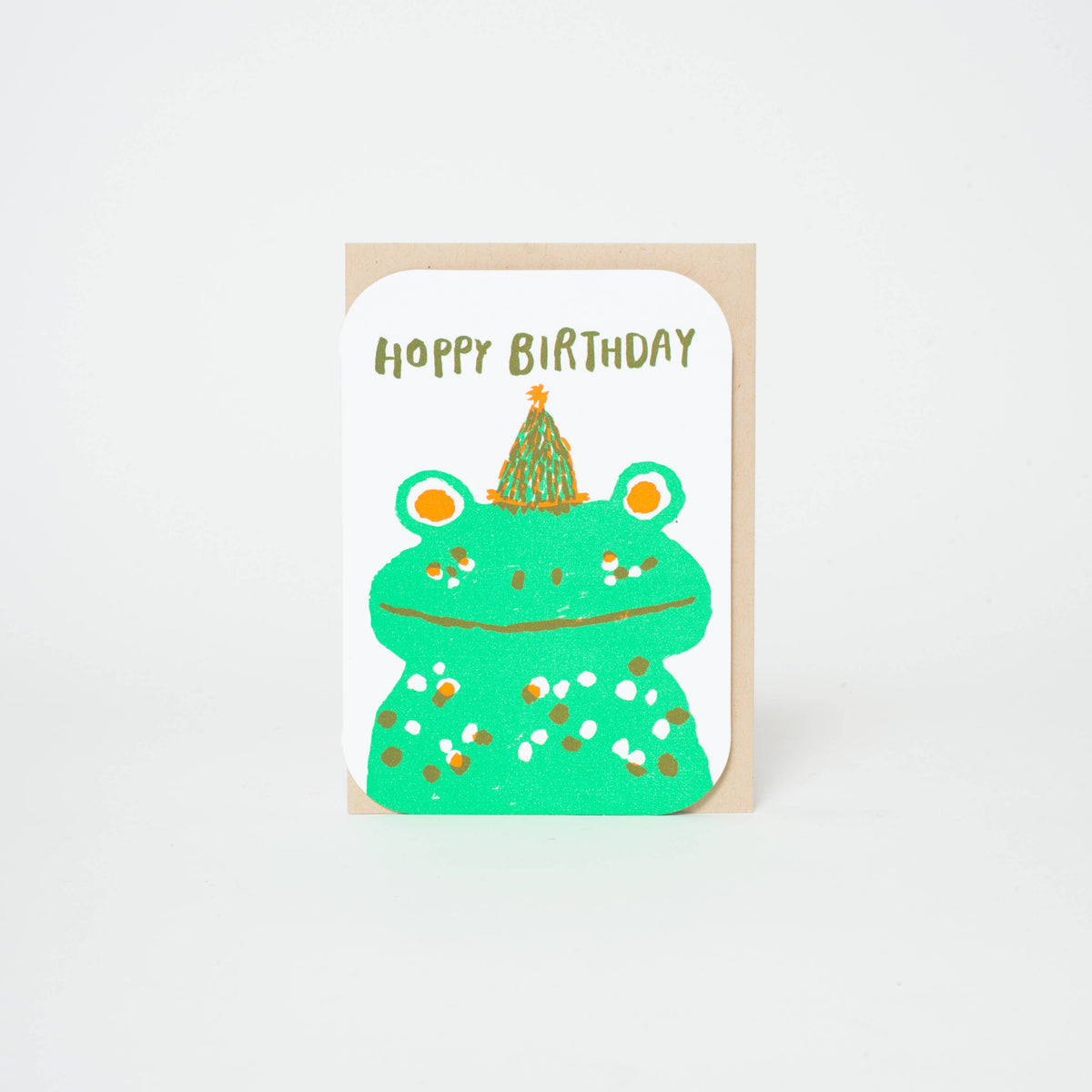 A square birthday card with a white front and kraft paper backing, featuring a green frog wearing a party hat and the words 'HAPPY BIRTHDAY' in olive green. The card is photographed on a clean white background. by Egg Press